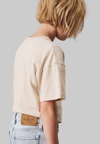 Beige bavlněné tričko s krátkým rukávem, s kulatým výstřihem a hnědou koženou nášivkou s nápisem "Calvin Klein Jeans" na zadním lemu.