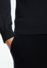 Navy-blauer Strickpullover mit gerippten Bündchen und Saum, mit glatter Textur und modernem Schnitt, teilweise gezeigt mit einer Hand in der Tasche.