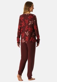Vrouw die blootsvoets staat, met een bordeauxrode lange mouwen top met rode en roze bloemenpatronen en bijpassende bordeauxrode broek, zijwaarts gekeerd.