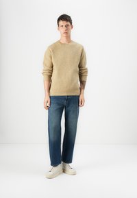 Suéter bege de malha com textura canelada, combinado com jeans de perna reta azuis e ténis brancos, em pé contra um fundo branco liso.