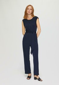 Navyblauwe jumpsuit met korte mouwen, een ronde halslijn en een taille met riem. Losvallende broekspijpen tot halverwege de kuit. Draagt men met zwarte haksandalen.