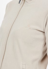 Giacca in pile beige con colletto alto, zip frontale e logo minimalista sul lato sinistro del petto. Texture morbida e design essenziale.