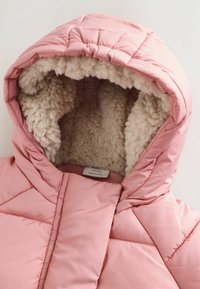 Vaaleanpunainen puffatakki, jossa on tyylitelty ruutukuvio, ja jossa on pehmeä, kermaisen värinen vuori hupussa ja kauluksessa. Tagi näkyy ulkopuolella.
