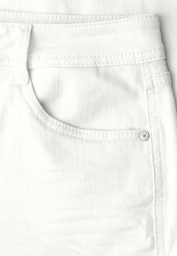 Gros plan sur la poche avant d'un jean blanc en denim avec des coutures visibles, une boucle de ceinture et un petit rivet métallique sur le bord de la poche.