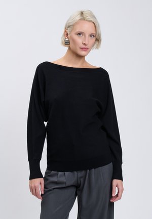 Blonde woman wearing large silver earrings, a loose black long-sleeve top, and gray pleated pants against a plain gray background.