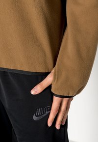 Hnědý fleecový pulovr s texturovanou tkaninou, s černým lemem na spodním okraji a rukávech. Černé logo Nike na tmavých kalhotách. Ruka odložená na látce.
