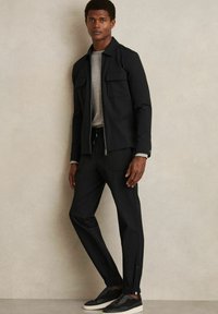 Black zip-up jacket with two front pockets, grey crew neck shirt, and tapered black pants with elastic cuffs. Black slip-on sneakers.