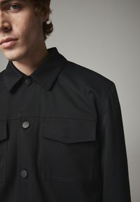 Sort arbejdsjakke lavet af struktureret stof med spidse kraver, trykknapper og to brystlommer med klapper.
