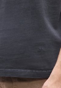 Κοντινό πλάνο της ραφής ενός σκούρου γκρι πουκαμίσου με μικρά κεντημένα αρχικά "MN" και εμφανές τμήμα του χεριού ενός ατόμου στη δεξιά πλευρά.