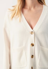 Crème cardigan gemaakt van zachte gebreide stof. Voorzien van een V-hals, vier grote bruine knopen en twee voorzakken met ribbeldetails.