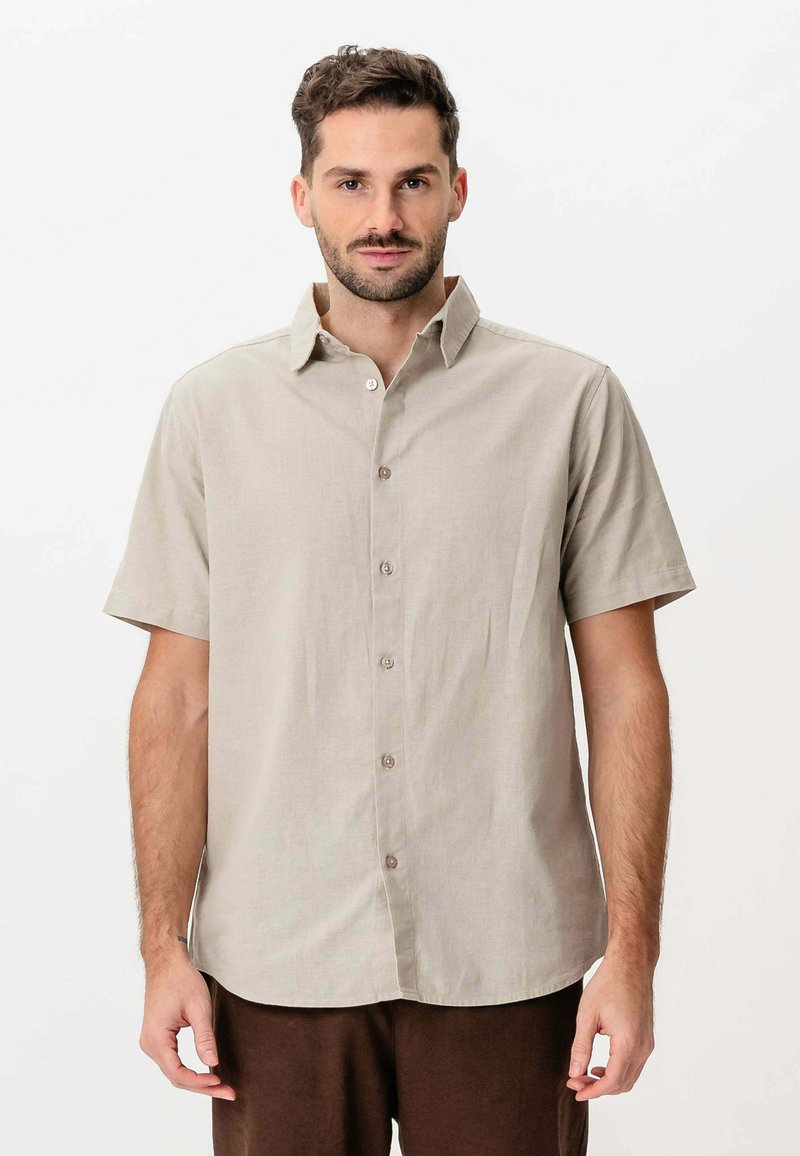 Mand med kort hår og skæg iført enbeige kortærmet skjorte med knapper og mørkebrune bukser, der står foran en ensfarvet hvid baggrund.