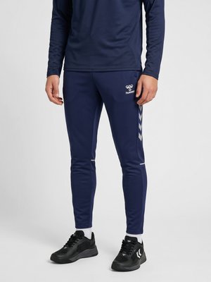 Mand iført marineblå sportbukser med hvide chevronstriber og sorte sneakers, stående mod en ensfarvet baggrund.