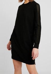 Abito maglione nero con vestibilità comoda. Presenta una texture a coste sulle maniche, spacco laterale e scollo rotondo. Tessuto in maglia morbida.