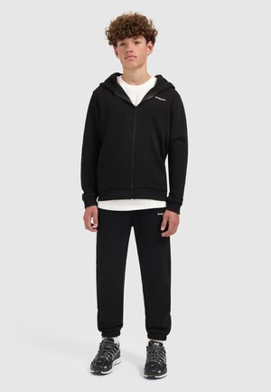 Tiener met krullend haar in een zwarte zip-up hoodie, bijpassende zwarte trainingsbroek, wit T-shirt en zwarte sportschoenen, staand op een witte vloer.
