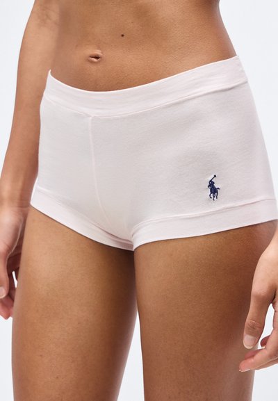 Κλείσιμο σε πρόσωπο που φοράει ανοιχτό ροζ, εφαρμοστά βερμούδακια με ένα μικρό σκούρο μπλε κεντημένο λογότυπο στο αριστερό ισχίο.