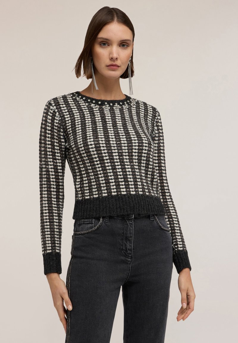 Gestrickter schwarz-weißer Pullover mit gestreiftem Muster, rundem Halsausschnitt und geripptem Saum. Kombiniert mit dunklen Jeans, die eine figurnahe Passform zeigen.