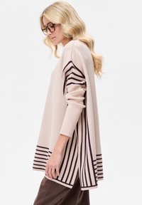 Beige oversized trui met lange mouwen, voorzien van verticale donkerbruine strepen aan de zijkanten en een effen onderste zoom. Zachte textuur.