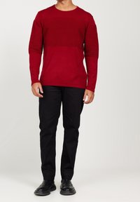 Maglione rosso a maniche lunghe con texture a costine nella parte superiore e parte inferiore liscia, abbinato a pantaloni neri e scarpe.