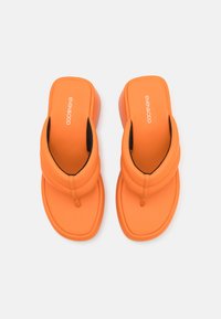 Oransje sandaler med tykk såle og mykt overdell, med en tåpost og en glatt tekstur. Logo trykt på den indre remmen.