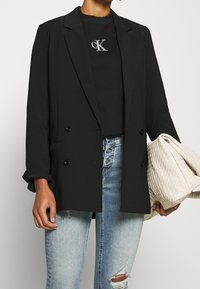 Schwarzer, doppelt geknöpfter Blazer mit hochgekrempelten Ärmeln, kombiniert mit einem schwarzen Rundhalsshirt mit silbernem Logo, abgenutzten hellblauen Jeans und einer strukturierten beigen Tasche.