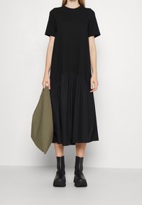 Schwarzes Midi-Kleid mit kurzen Ärmeln, gerafftem unteren Teil und weicher Textur. Trägt dazu klobige schwarze Stiefel und hält eine olivgrüne Tasche.