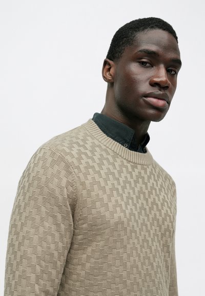 Jauns vīrietis ar tumšu ādu valkā smilškrāsas tekstūru adītu džemperi virs melnas krekla ar apkakli, nedaudz skatoties uz sāniem uz vienkārša fona.