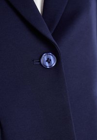 Blazer navy con colletto a vela, tessuto testurizzato e dettaglio a bottone blu tondo con quattro fori.