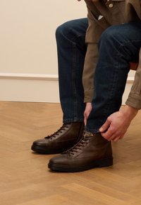 Bruine leren enkellaarzen met vetersluiting, ronde neus en subtiele textuurafwerking. Draag ze met donkerblauwe jeans, zittend op houten vloerdelen.