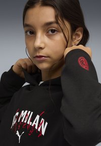 Sweat à capuche noir avec l'inscription "AC MILAN" en blanc, agrémenté de flammes rouges. Un petit logo rouge est présent sur la manche. Texture de tissu lisse.