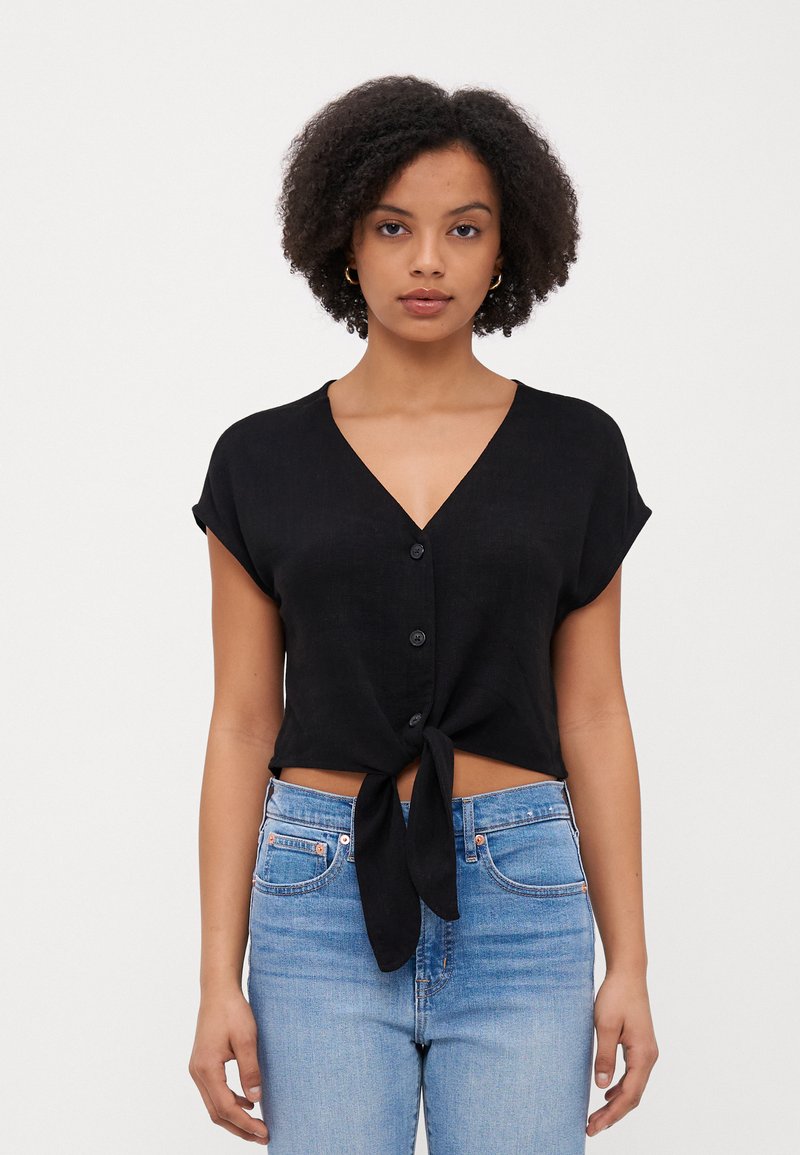 Maglietta nera corta con scollatura a V, maniche corte, chiusura con bottoni e un nodo all'orlo. Indossata sopra jeans azzurri chiari.