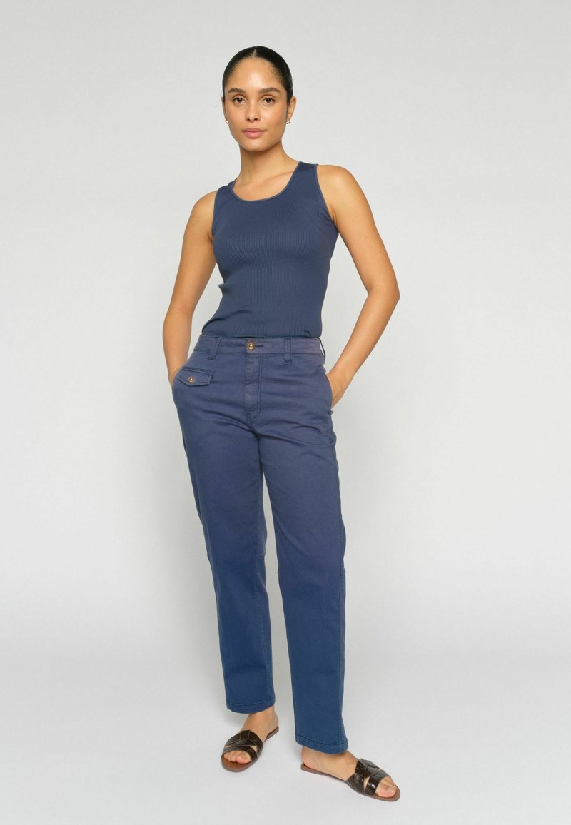 Femme debout les mains dans les poches d'un pantalon bleu, portant un haut sans manches bleu marine et des sandales noires à bout ouvert, sur un fond clair uni.