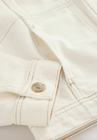 Gros plan sur le poignet d'une veste en denim beige avec un bouton beige et des détails de couture visibles.