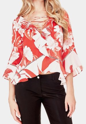Kobieta ubrana w czerwoną i białą kwiecistą bluzkę kopertową z falbankami i rękawami dzwonkowymi, w połączeniu z czarnymi spodniami.