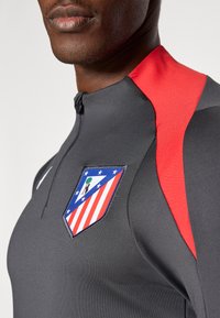 Szürke athletic póló piros kiemelésekkel, félig cipzáras gallérral és kék, fehér, piros mintázatú logóval. Sima textúra, testhezálló fazon.