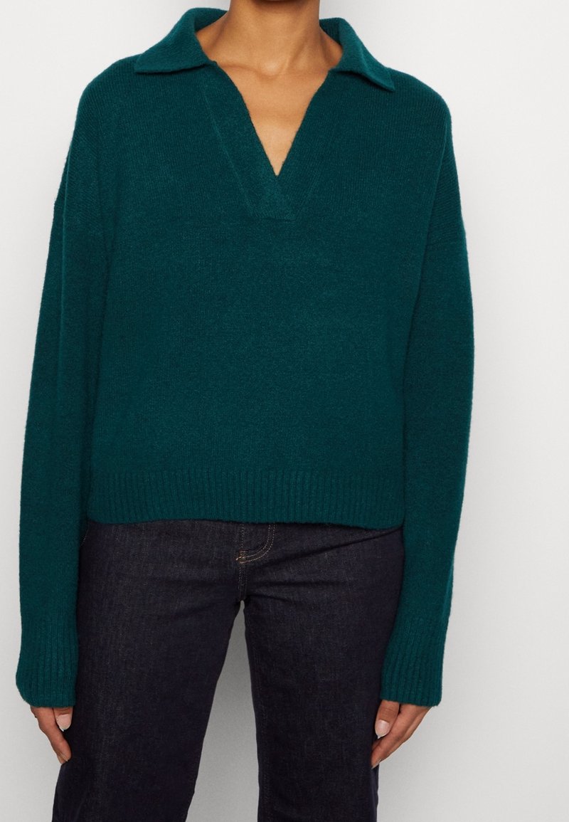 Personne portant un pull en maille vert foncé à manches longues avec un col et un profond décolleté en V, associé à un jean foncé.