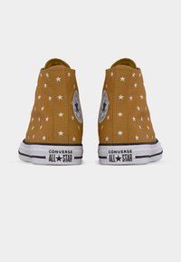 Υψηλά καμβά παπούτσια σε μουσταρδί χρώμα με λευκό μοτίβο αστεριού, με μαύρες λεπτομέρειες και κυκλικό λογότυπο στο εξωτερικό μέρος.