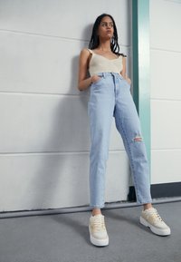 Ljusblå högmidjade jeans med ett hål vid knäet, kombinerat med en krämfärgad ribbad tanktop och beige sneakers med vita sulor.