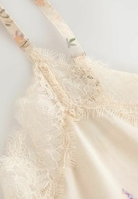 Camisole avec dentelle, ornée de bretelles à motif fleuri, en tissu beige doux et avec des détails en dentelle complexes le long de l'encolure et de l'ourlet.