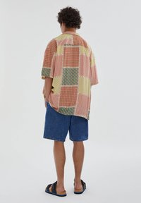 Camicia patchwork a maniche corte, colorata con motivi rosa, verdi e gialli, abbinata a pantaloncini di jeans e sandali neri.