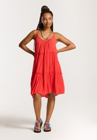 Rochie roșie, fără mâneci, cu un design în straturi și o margine ondulată. Are bretele subțiri și un material moale, texturat. O pereche de sandale colorate.