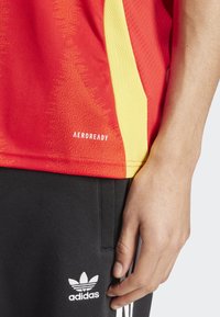 Crvena i žuta atletska majica od teksturiranog materijala s mrežastim detaljima; s oznakom "AEROREADY" na rubu; uparen s crnim hlačama koje imaju logo.