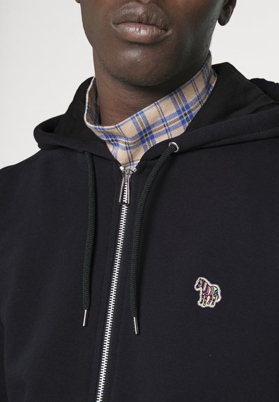 Marinblå zip-up hoodie med svart huva, med en färgglad zebra-logotyp på bröstet och en rutigt krage i blått, beige och vitt.