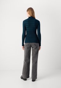 Pull en col roulé teal avec une texture côtelée et des rayures horizontales dans le dos, associé à un jean bleu gris ample taille haute et des chaussures noires.