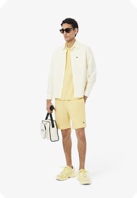 Outfit informale con polo gialla e pantaloni corti abbinati, completato da una camicia a righe con bottoni e una borsa bianca con dettagli neri.