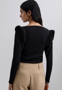 Woman with long dark hair wearing a textured black long-sleeve top with puffed shoulders and beige high-waisted pants, facing sideways.