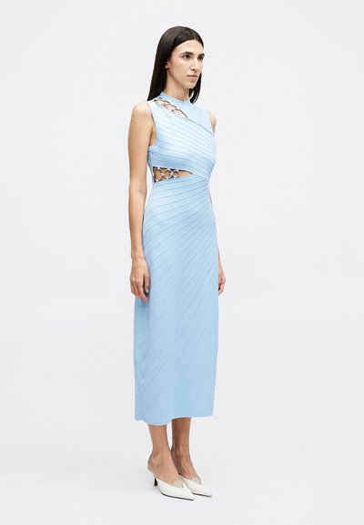 Nő áll, világoskék ujjatlan midi ruhát visel, amelyen átlós vonalak és kivágások vannak a vállánál és a deréknál, fehér magassarkúval párosítva.