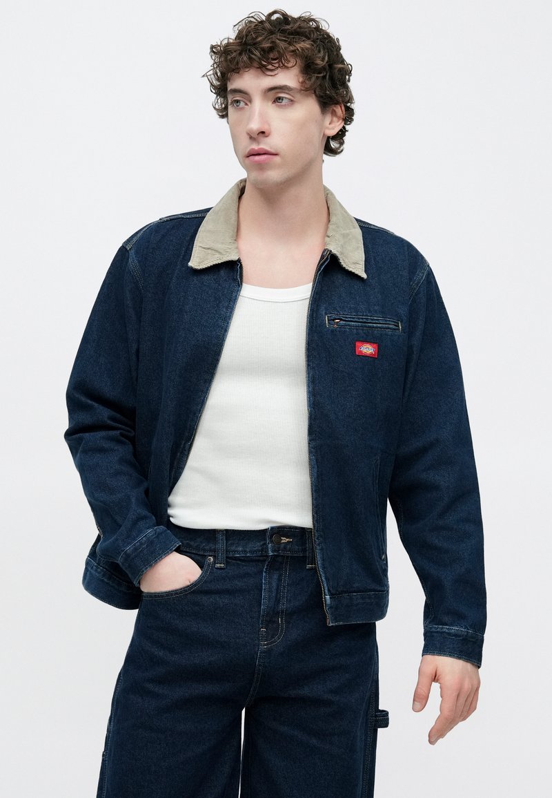 Veste en denim bleu foncé avec col en velours côtelé beige, poche avant et écusson logo rouge. Portée sur un débardeur blanc et un pantalon en denim foncé.