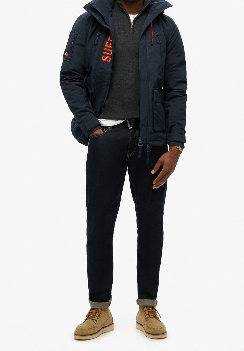 Homme portant une veste matelassée bleu marine avec un logo orange, un jean foncé, un pull gris et des bottes en daim beige, debout avec une main dans la poche.