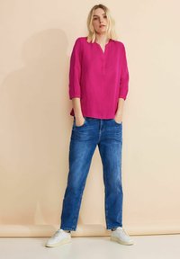 Blusa fucsia con popover, a tre quarti, vestibilità rilassata e una piccola scollatura, abbinata a jeans blu a gamba dritta e scarpe bianche.