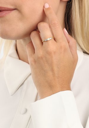Anello in oro giallo con tre gemme incastonate, finitura lucida, montato su una fascia delicata. Mostrato a mano vicino al viso.